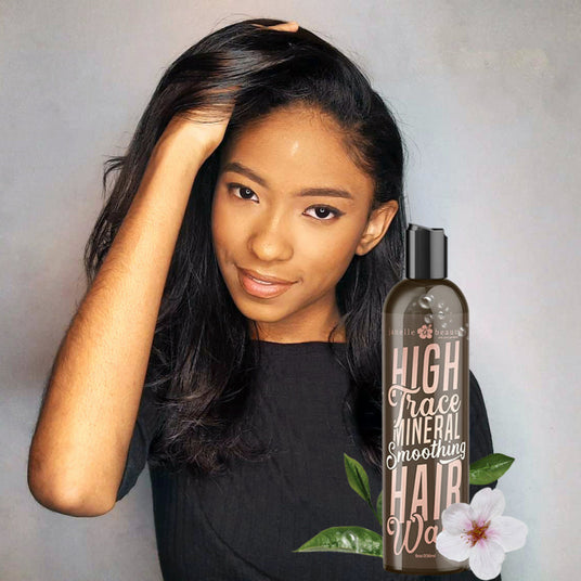Woman with long black hair holding a bottle of 'High Trace Mineral Smoothing Hair Wax' against a neutral background.